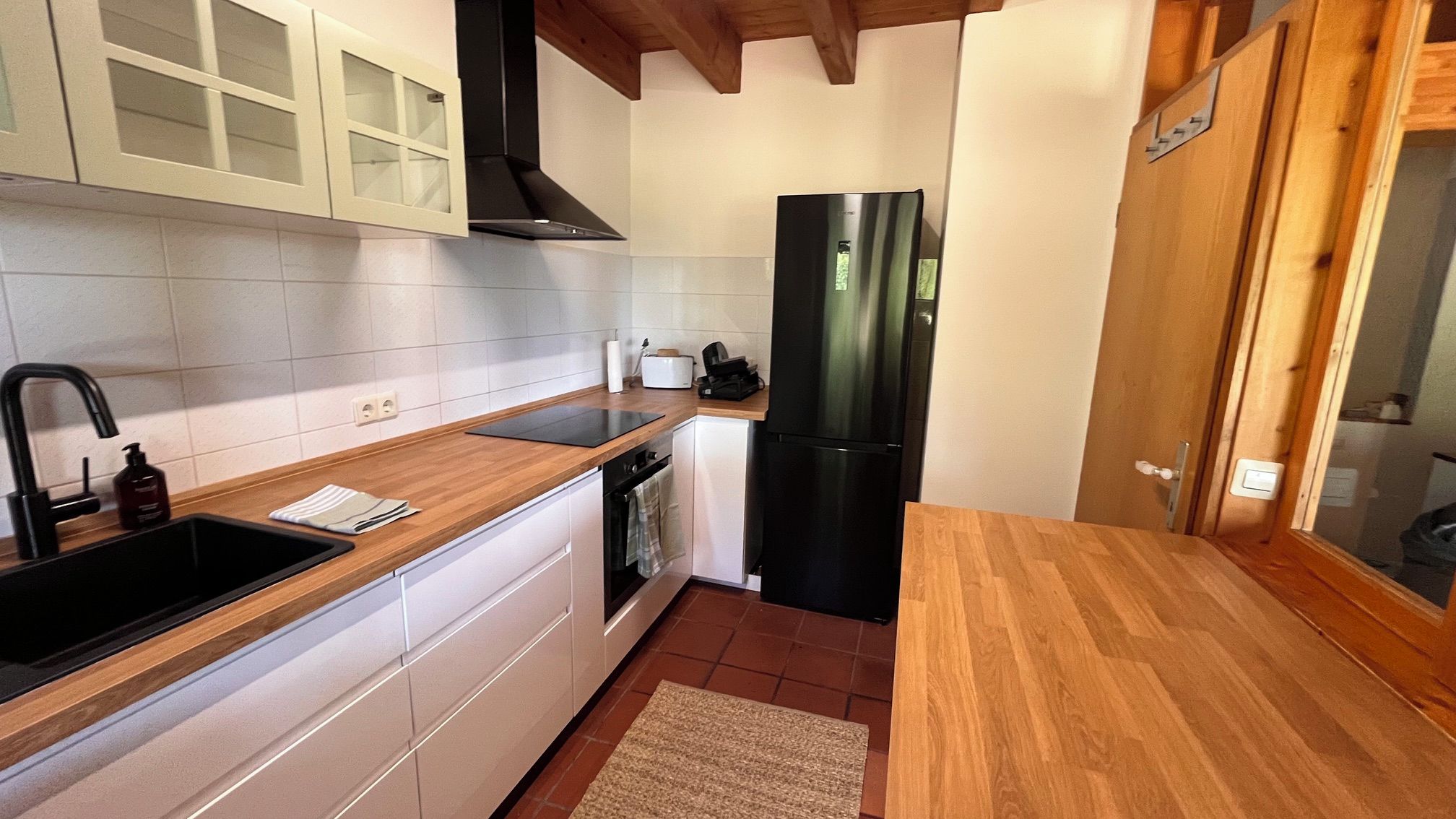Eine kleine Küche mit weißen Schränken, einem schwarzen Kühlschrank und einer Holz Arbeitsplatte.
