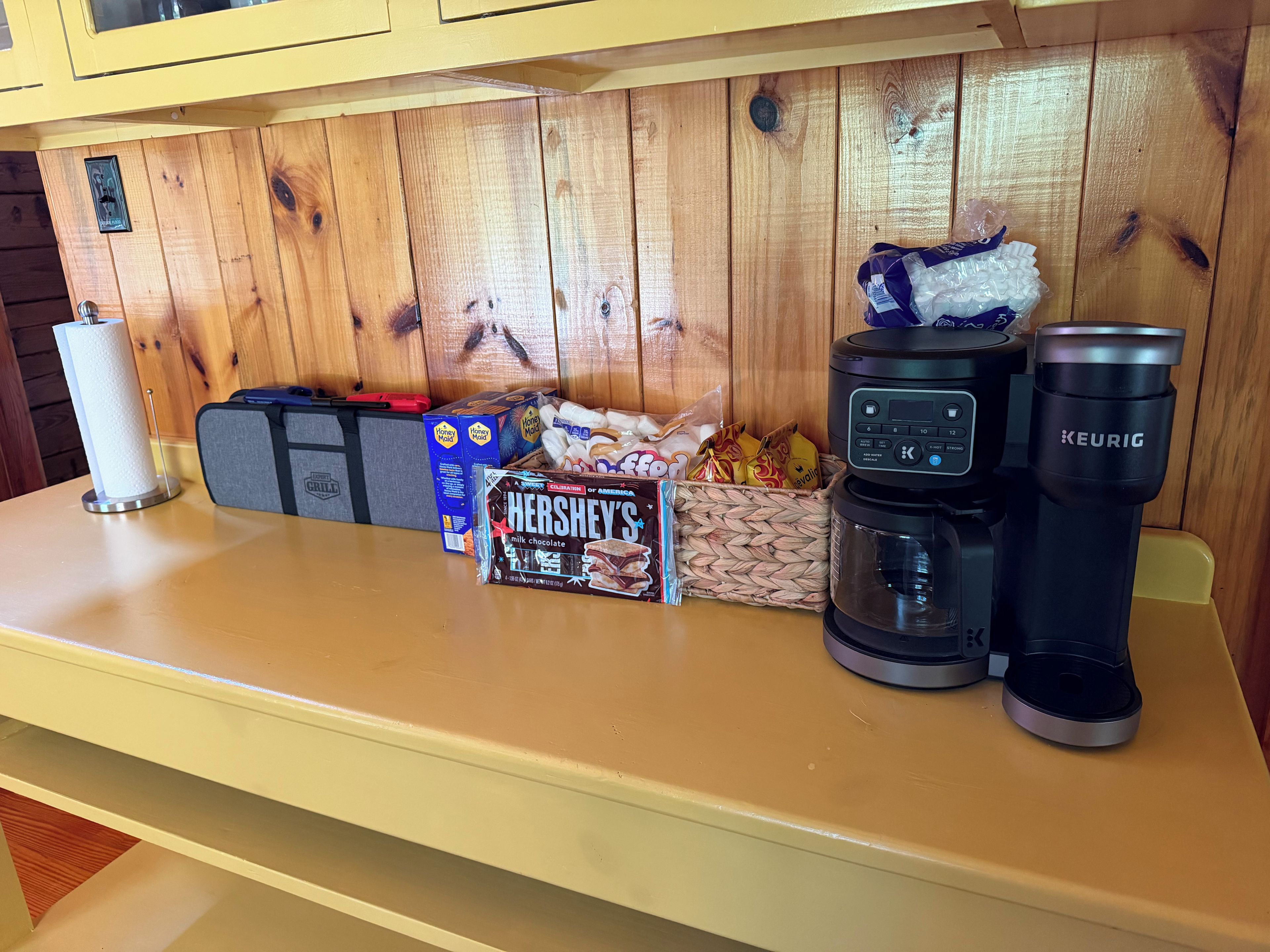 Eine Küchenarbeitsplatte mit einer Kaffeemaschine, einer Keurig, einer Schachtel Hershey's Milchschokolade, einer Honey Maid Schachtel und einem Snackkorb.