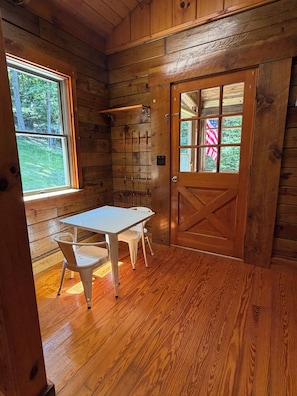 A wooden interior with a table and chairs, a door with an American flag, and a window with a view of trees.