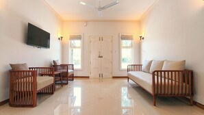 A spacious living room with a flat-screen TV mounted on the wall, a ceiling fan, and a large door with windows. - Estate IV By Zomestay (Vythiri)