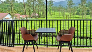 A balcony with two chairs and a table, overlooking a lush green landscape with tea plantations and houses. - Estate IV By Zomestay (Vythiri)