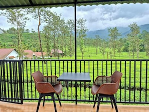 A balcony with two chairs and a table, overlooking a lush green landscape with tea plantations and houses. - Estate IV By Zomestay (Vythiri)