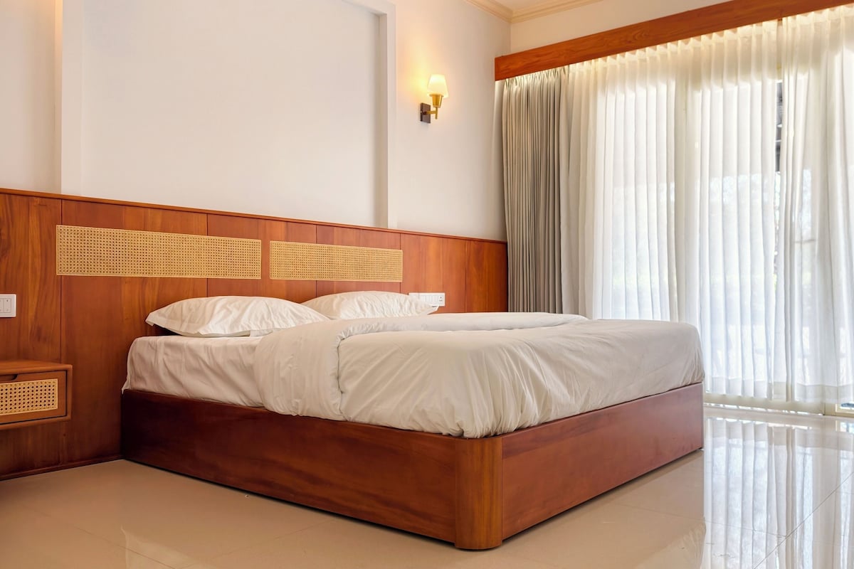 A hotel room with a wooden headboard, a bed with white bedding, a wall-mounted light, and sheer curtains.