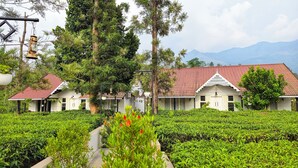 A building with a red-tiled roof and white walls, surrounded by lush greenery and a tea plantation. - Estate IV By Zomestay (Vythiri)
