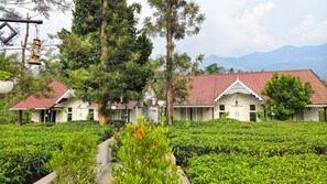 A building with a red-tiled roof and white walls, surrounded by lush greenery and a tea plantation. - Estate IV By Zomestay (Vythiri)