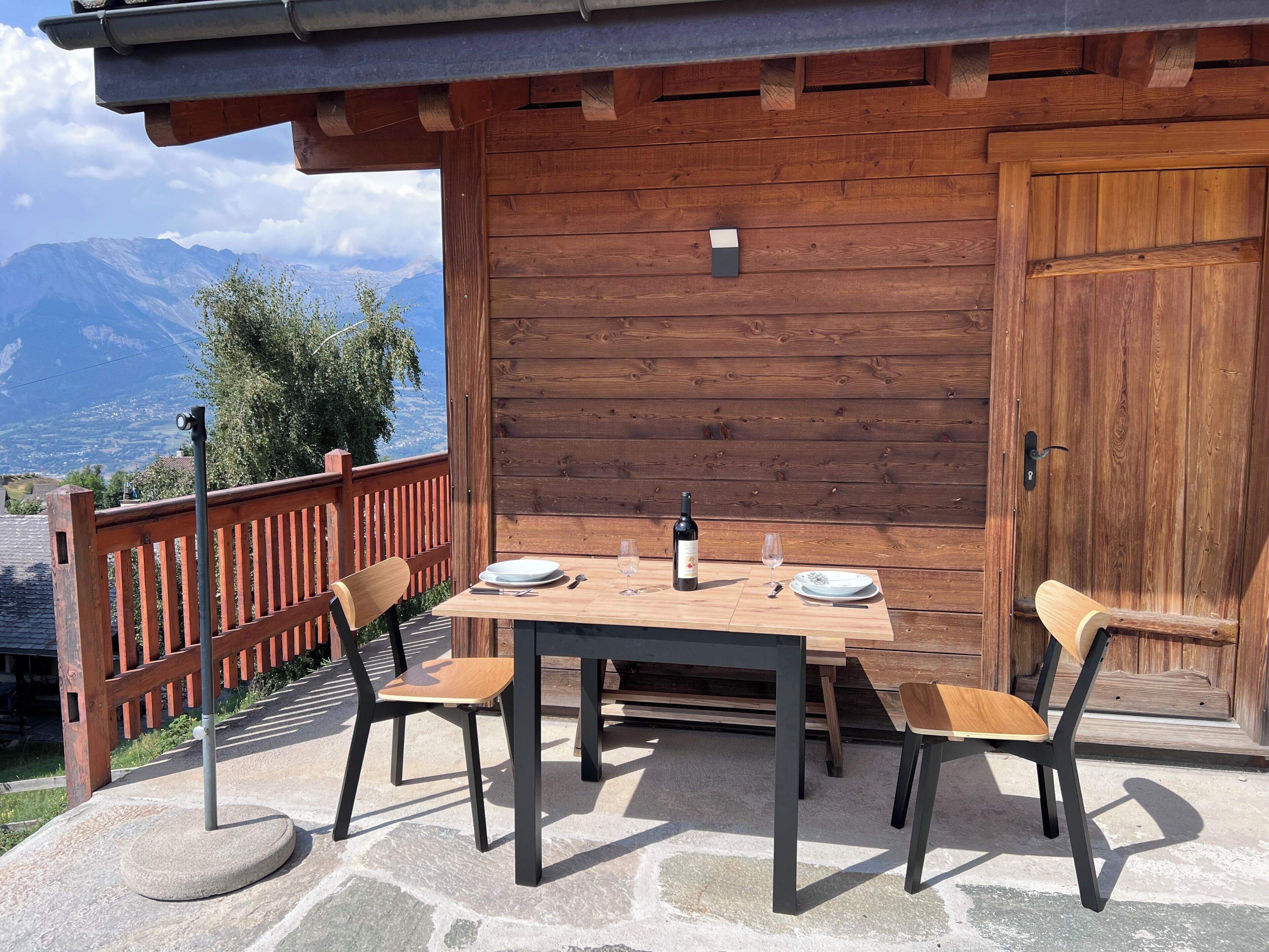 Un espace de repas extérieur en bois, avec une table dressée pour deux personnes, des chaises et une bouteille de vin.
