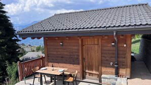 Een houten chalet met een stenen terras, een tafel met stoelen, en een kat die over het terras loopt.