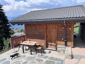 A wooden cabin with a stone patio, a table with chairs, and a cat walking on the patio. - Vrbo Property (Mont-Noble)