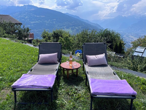 Two lounge chairs with purple cushions, a bottle of wine, and a glass on a small table, set against a backdrop of mountains and a clear sky.