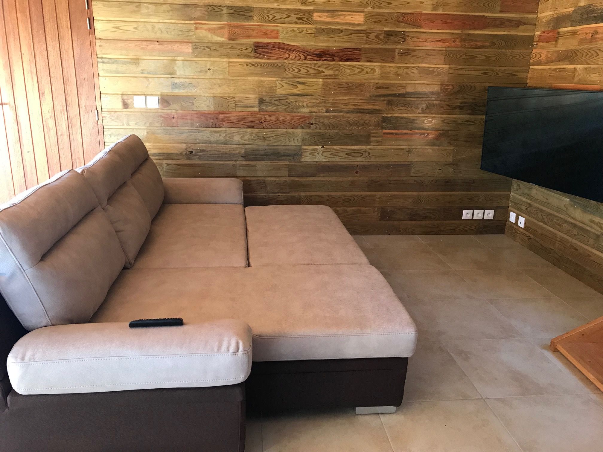Un sofá seccional color beige con un control remoto, ubicado en una habitación con pared de madera y un televisor de pantalla plana montado en la pared.