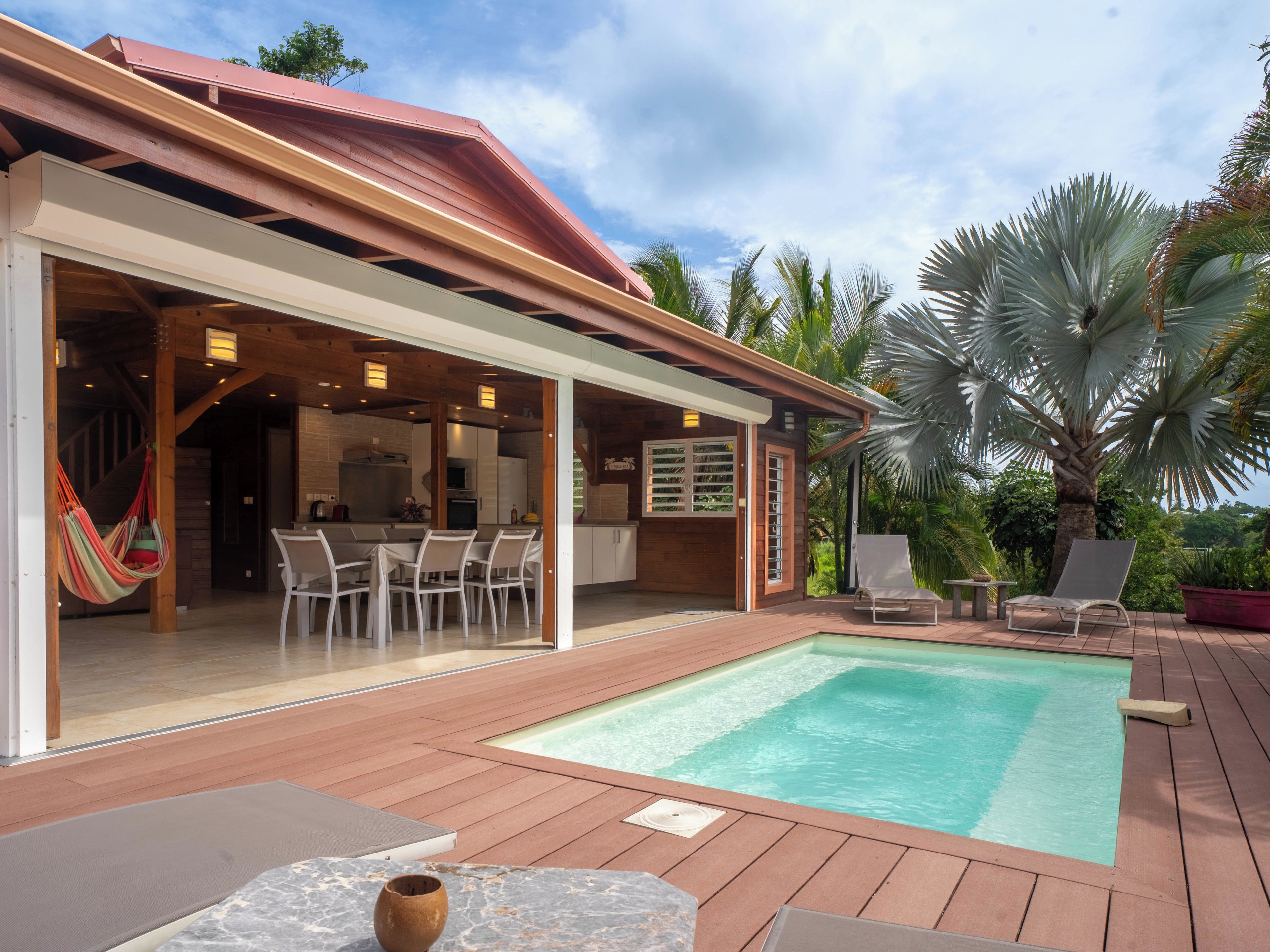 A pool area with a wooden deck, a dining area with chairs, and a hammock.