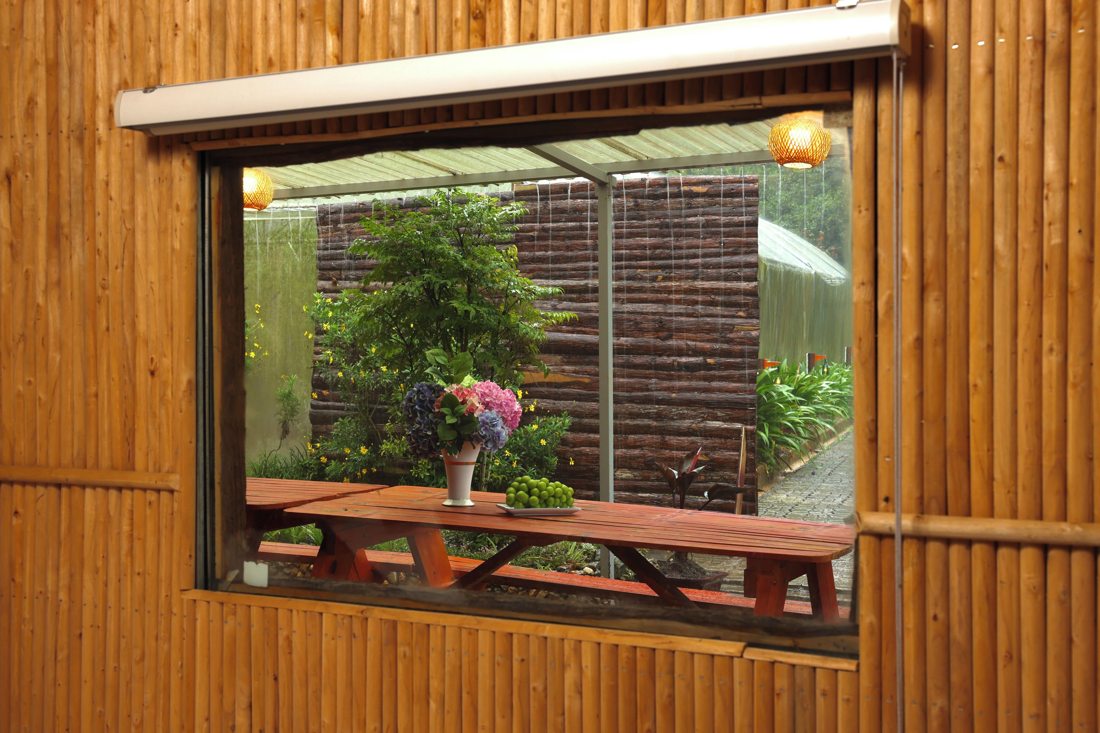 Una ventana con vistas a una pared de madera, una barandilla metálica y una mesa con un florero.