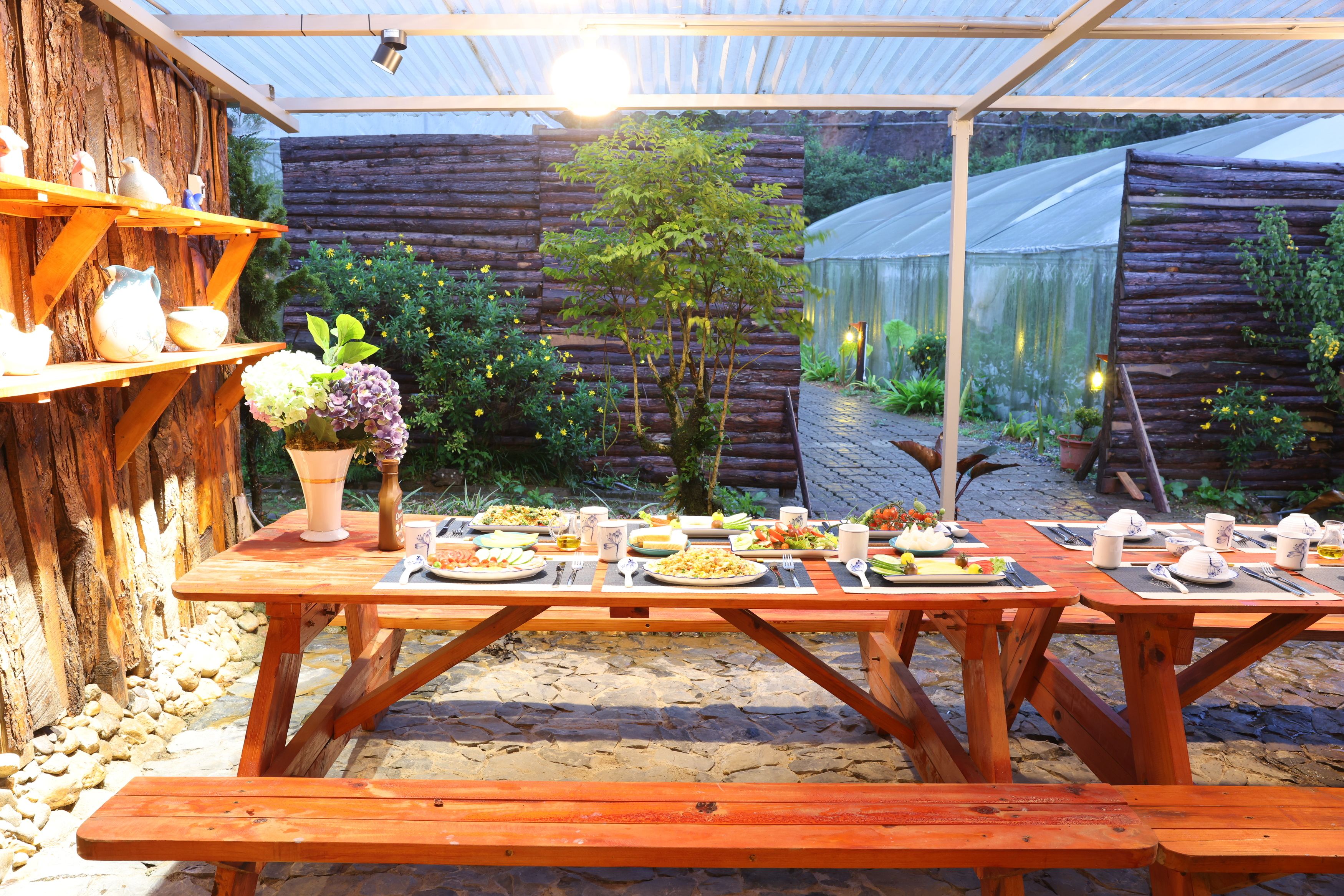 An outdoor dining area with wooden tables set for a meal, surrounded by a rustic wooden fence and a greenhouse structure.