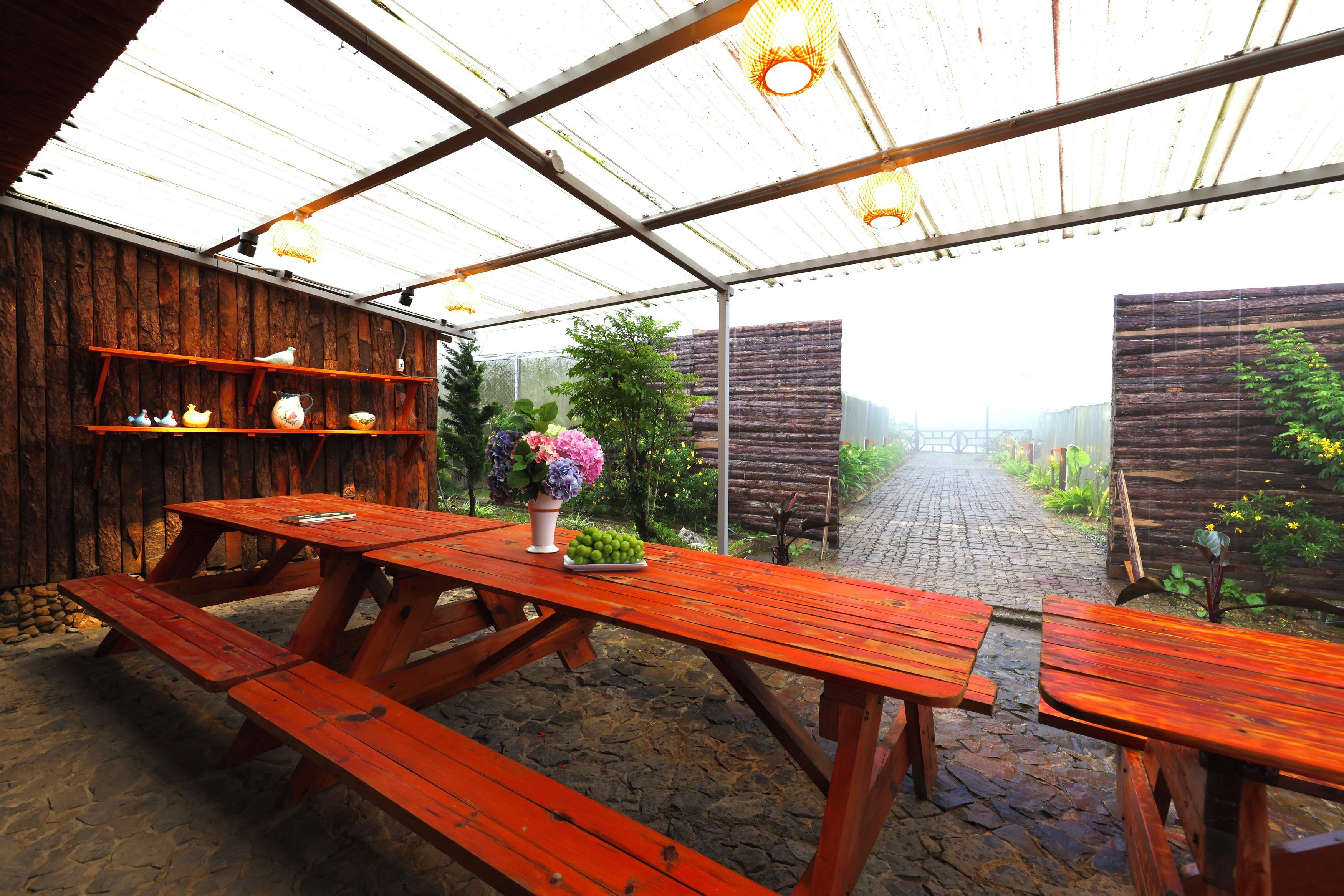 Un espacio de descanso al aire libre con bancos de madera y una mesa, rodeado por un rústico muro de madera y un sendero en el jardín.