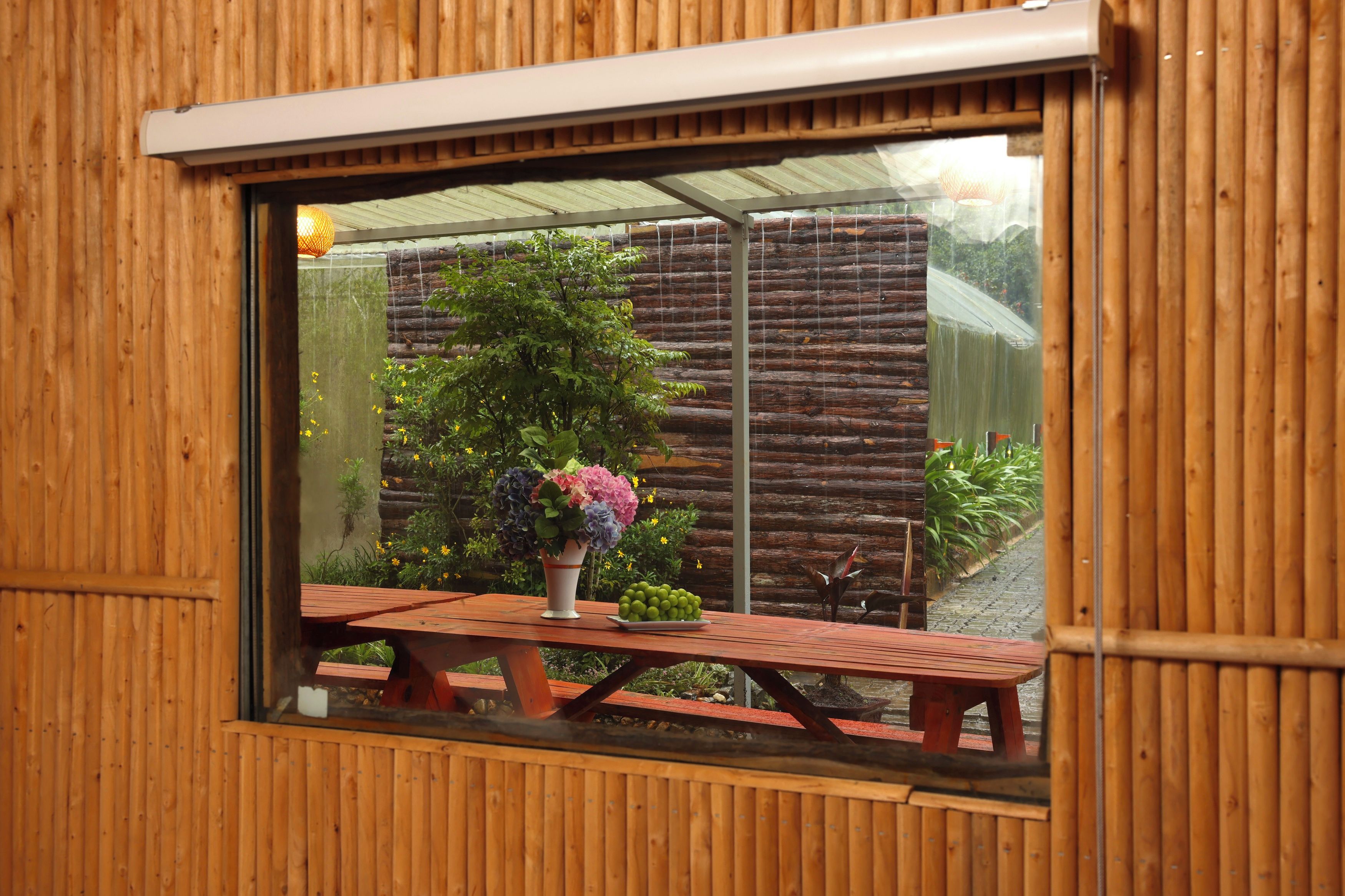 A wooden-framed window with a view of a garden and a picnic table with a vase of flowers.