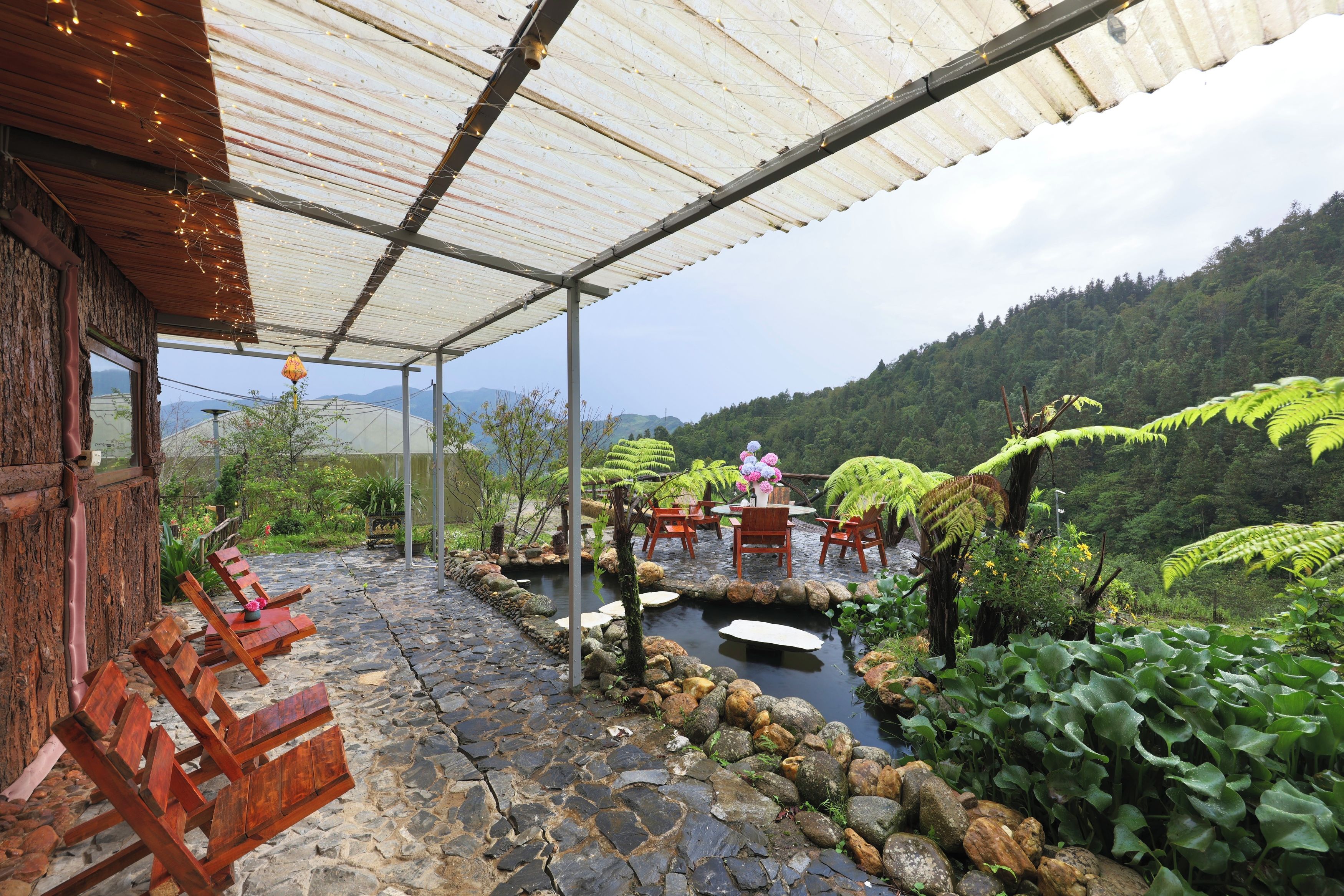 An outdoor seating area with stone flooring, wooden chairs, a small pond, and lush greenery.