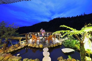 A wooden bridge over a reflective water body with a table and chairs, surrounded by rocks and greenery.