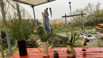 Un espacio al aire libre cubierto con una mesa de madera, plantas en macetas y un invernadero al fondo.