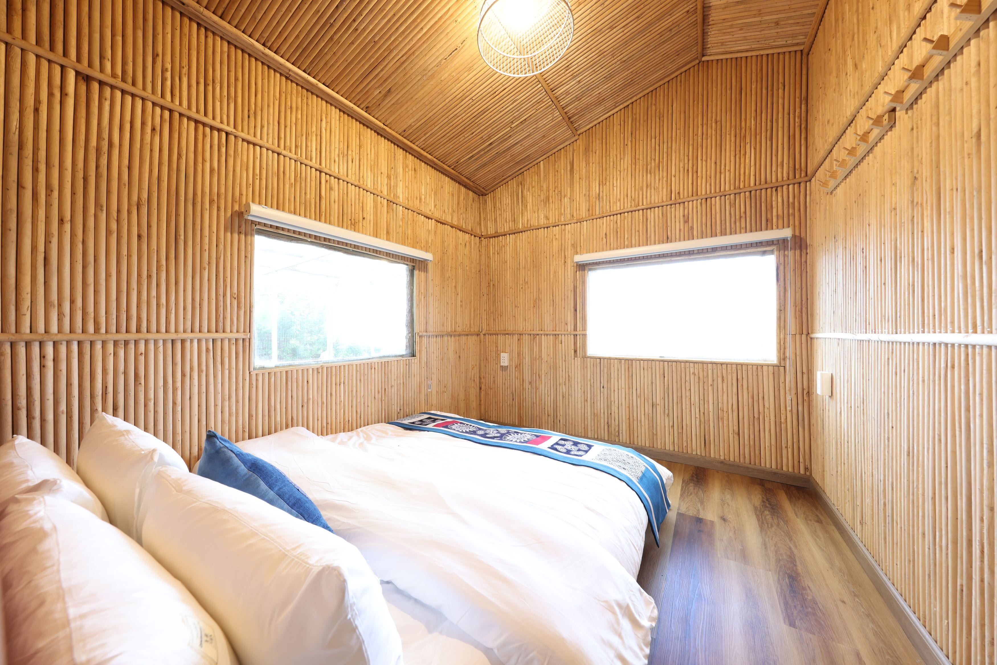 Una habitación con una cama, dos ventanas y paredes de madera.