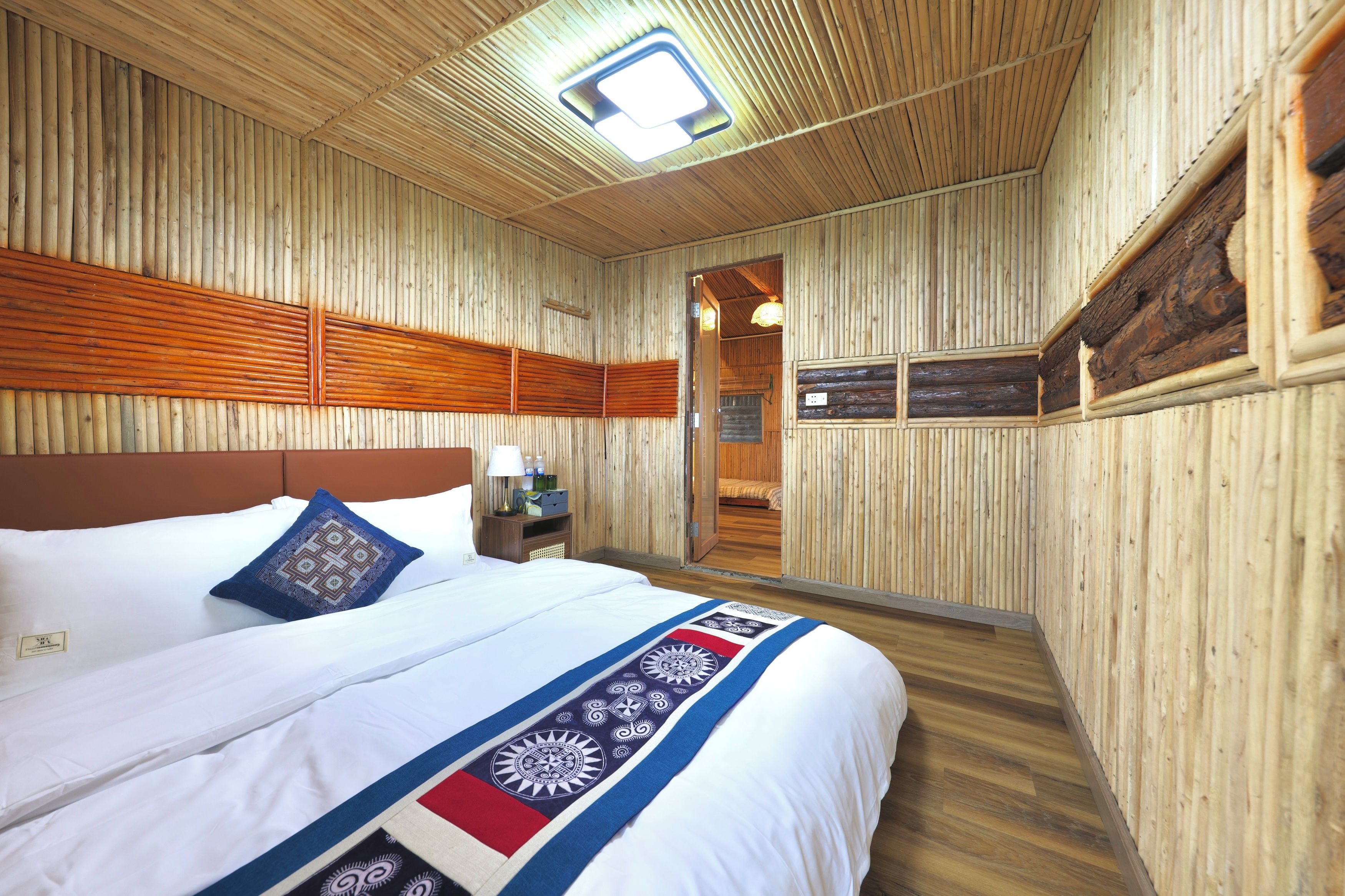 Un interior de madera con una cama, almohadas, una mesita de noche y una puerta que da a otra habitación.