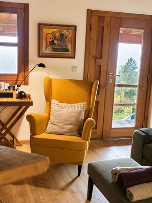 A yellow armchair with a white pillow, a wooden desk with a lamp, and a door leading to an outdoor area.