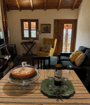 A cozy living room with a wooden ceiling, a dining table with a cake and wine glasses, a yellow armchair, and a sofa with cushions.