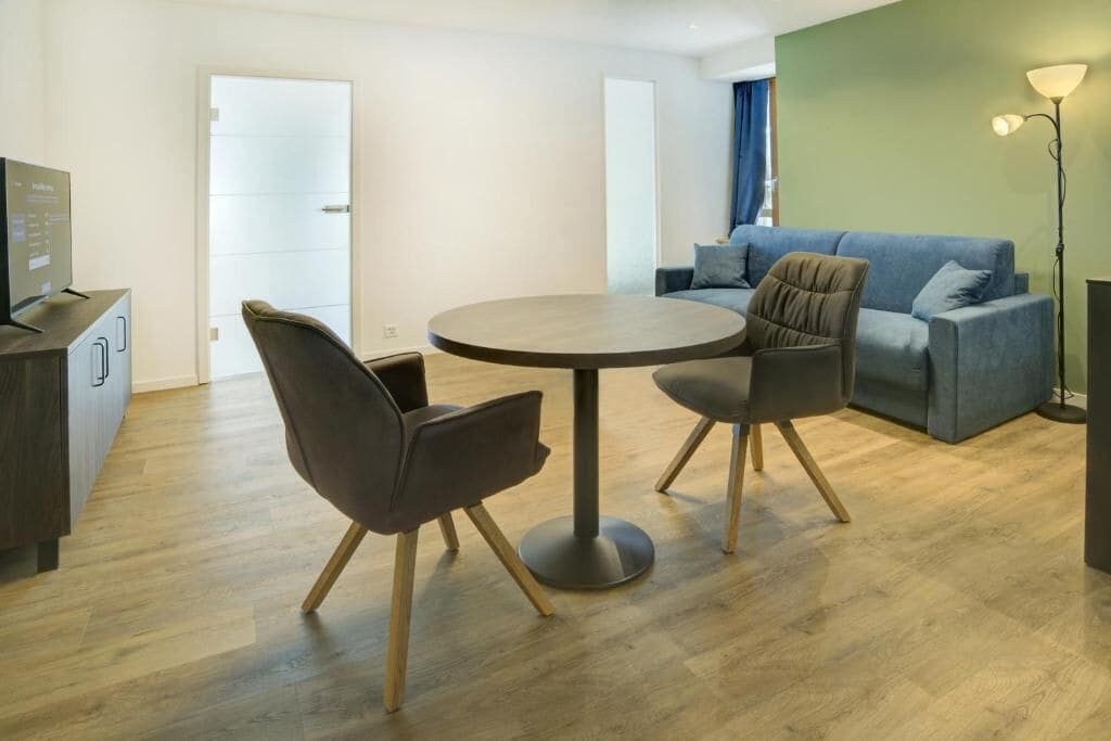 Ein modernes Wohnzimmer mit rundem Tisch und zwei Stühlen, einem Sofa, einem Fernseher und einer Lampe.