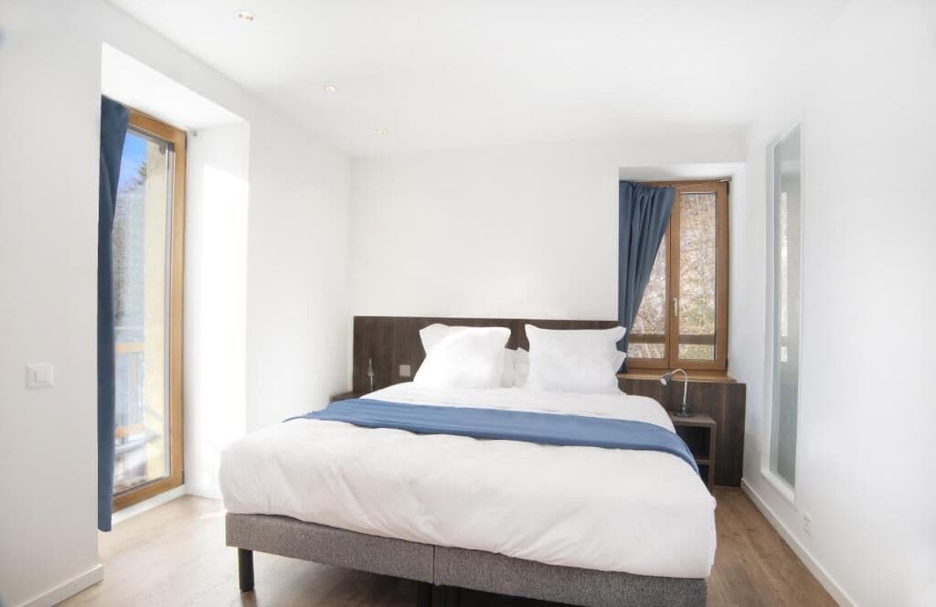Un dormitorio con cama, mesita de noche, ventana con cortinas y puerta que da a otra habitación.