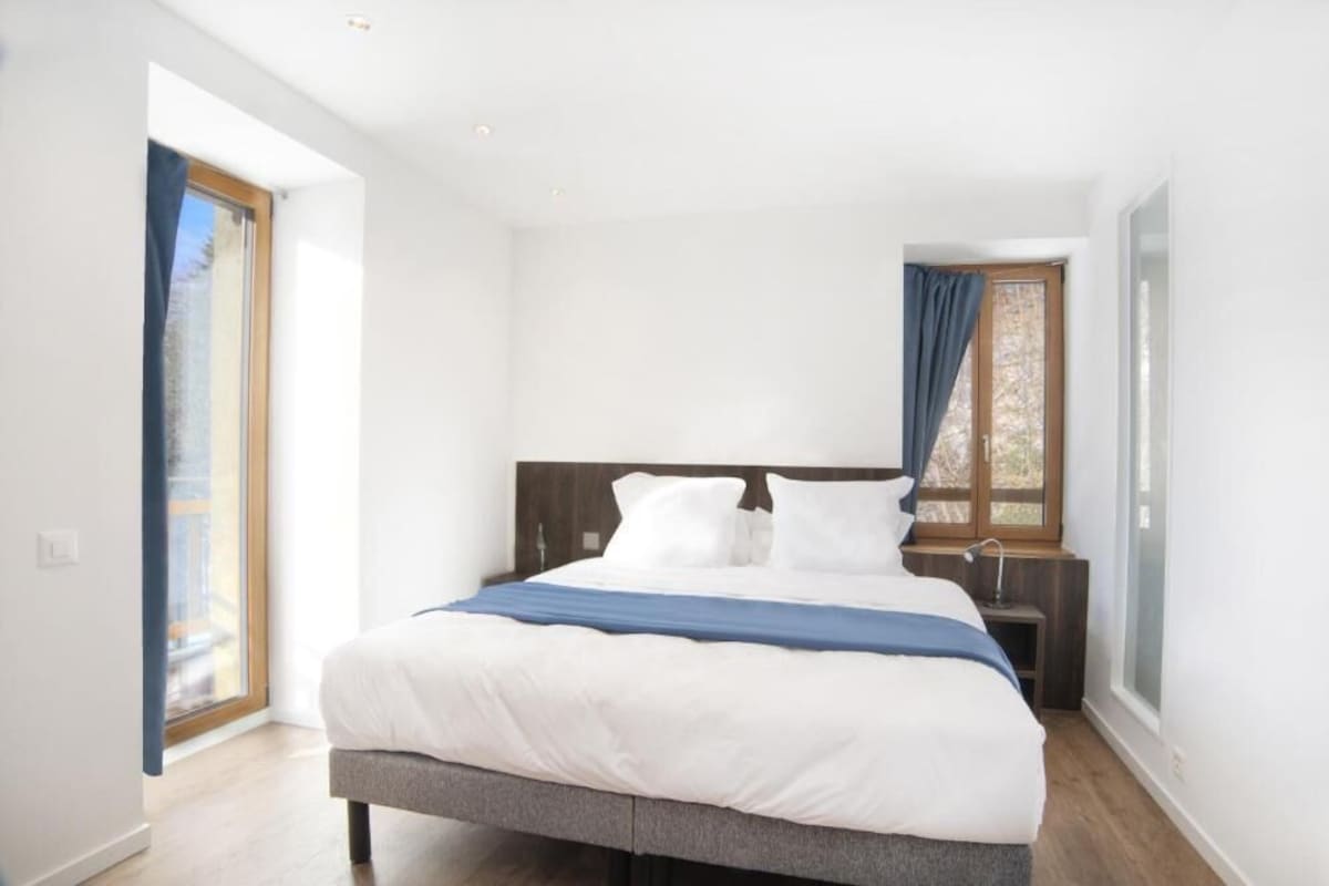 Quarto com cama, mesa de cabeceira, janela com cortinas e porta que dá para outro cômodo.