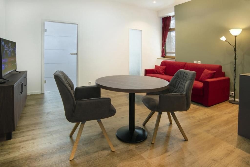 Un salon moderne avec une table ronde et quatre chaises, un ensemble de canapés rouge, un téléviseur posé sur un meuble et un lampadaire.