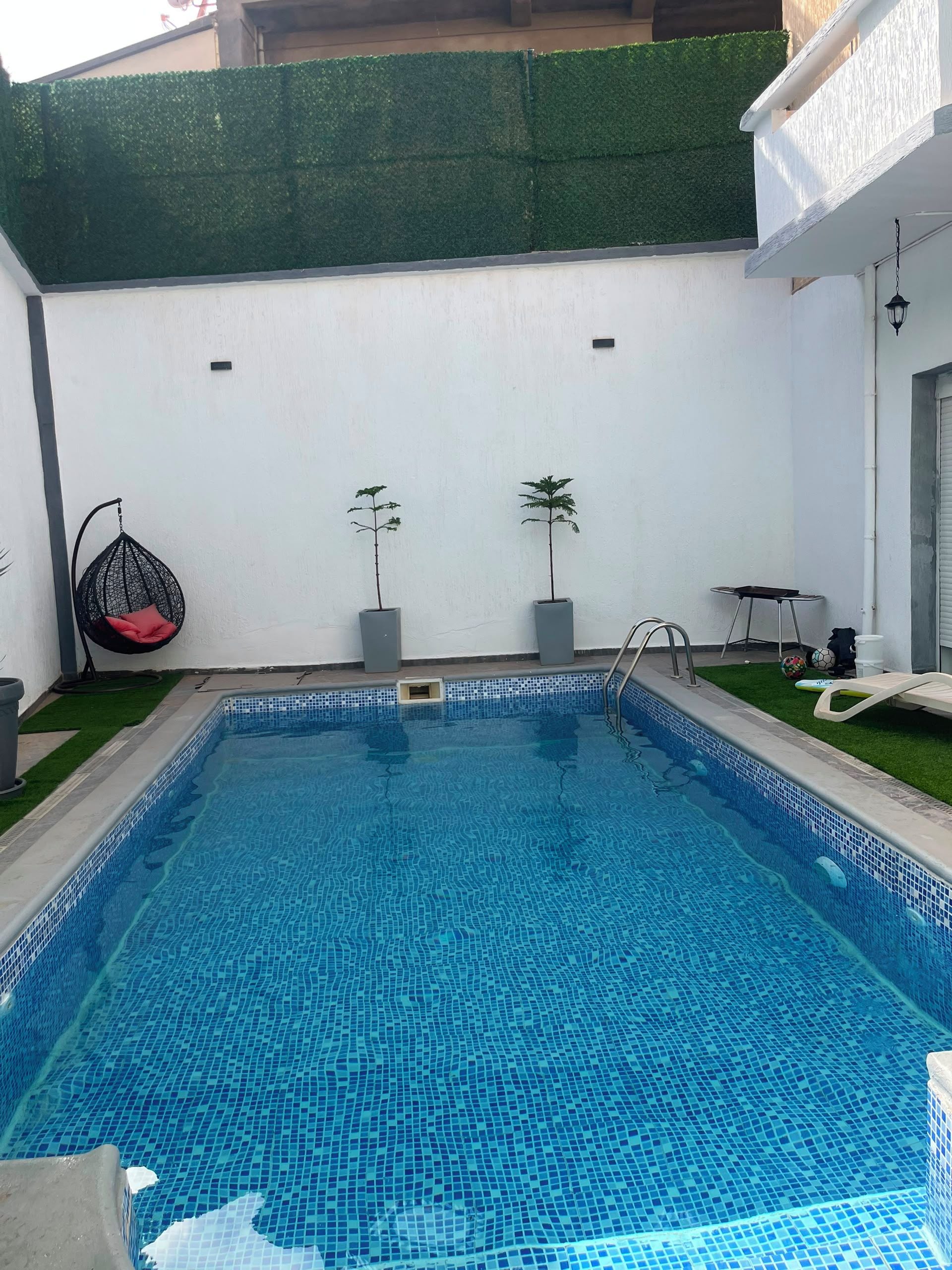 A rectangular swimming pool with a tiled edge, surrounded by a white wall with a green hedge. There are two potted plants, a hanging chair, and a small table with chairs.