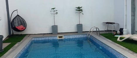 A rectangular swimming pool with a tiled edge, surrounded by a white wall with a green hedge. There are two potted plants, a hanging chair, and a small table with chairs.