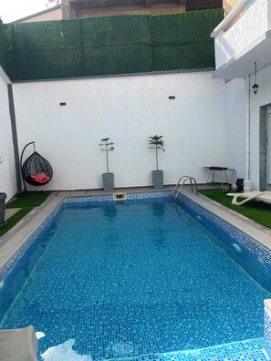 A rectangular swimming pool with a tiled edge, surrounded by a white wall with a green hedge. There are two potted plants, a hanging chair, and a small table with chairs.