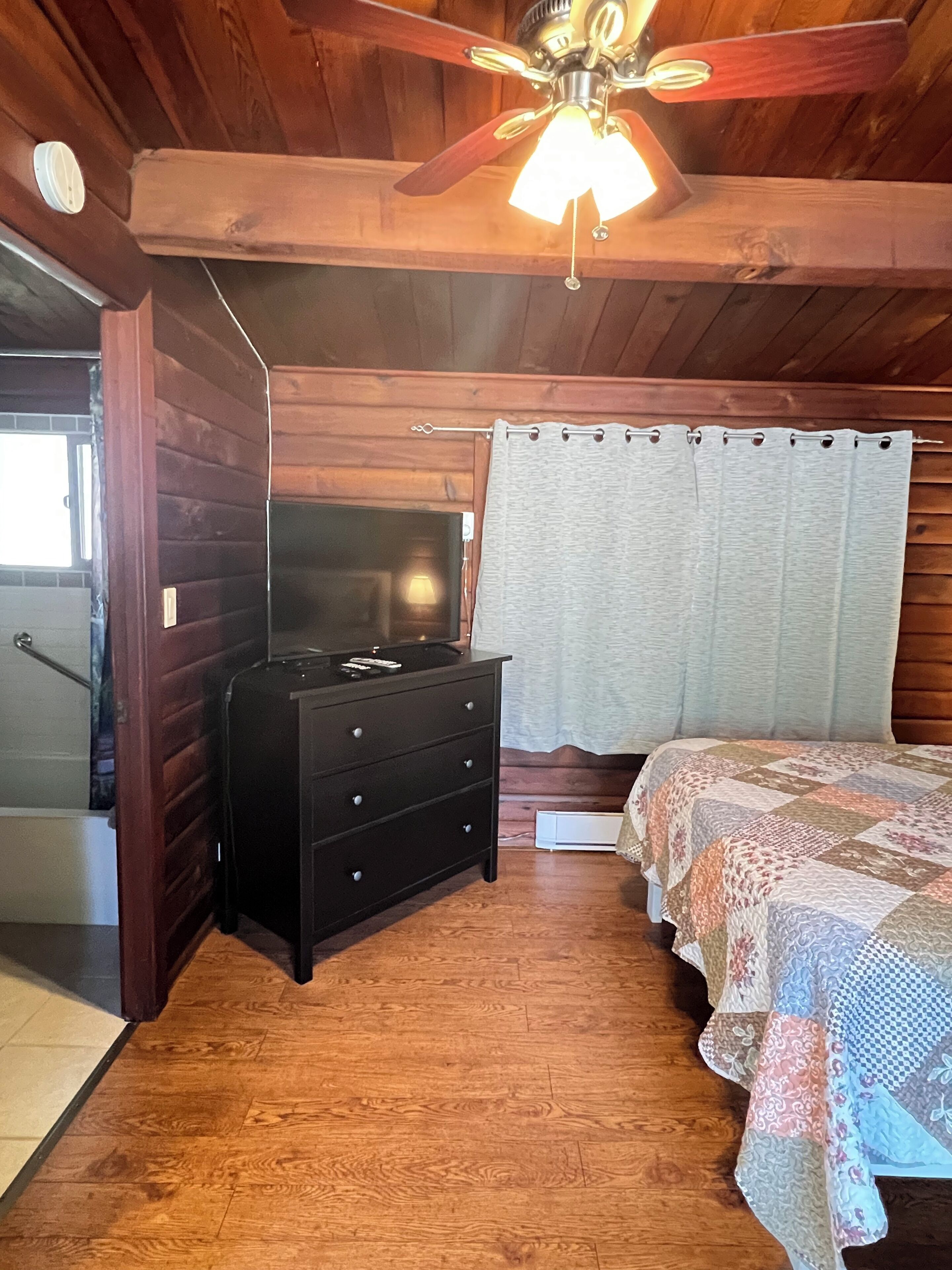 A wooden-paneled room with a ceiling fan, a bed with a patterned bedspread, a dresser, and a window with curtains.