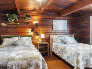 A cabin-style room with two beds, wooden walls, a hanging plant, a lamp, and a painting.