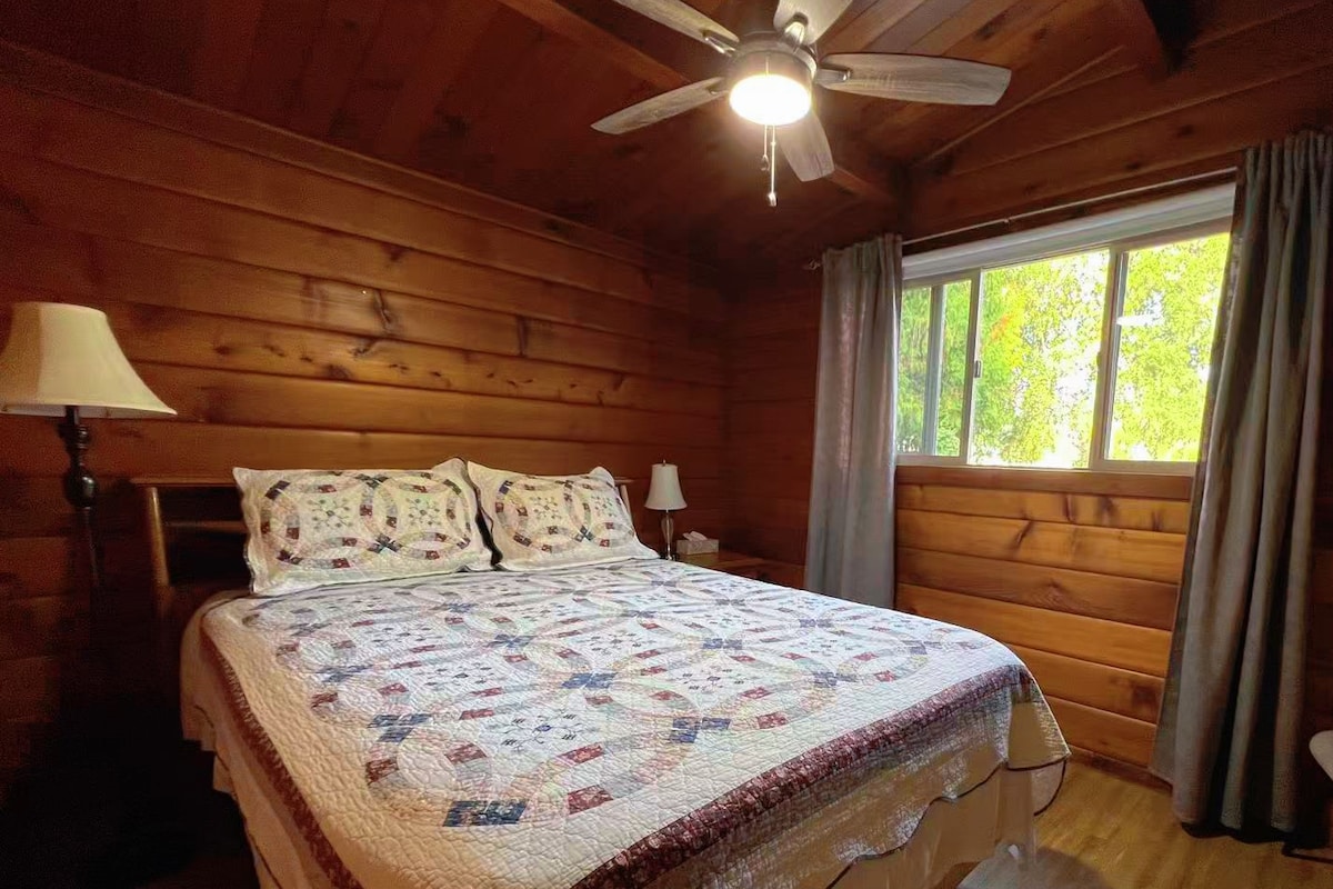 Une chambre dans une cabane en bois, avec un lit, un ventilateur de plafond, une fenêtre avec des rideaux et une lampe.