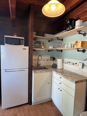 A compact kitchen with a white refrigerator, a microwave, a toaster, and a sink. There are wooden shelves above the counter holding various kitchen items.