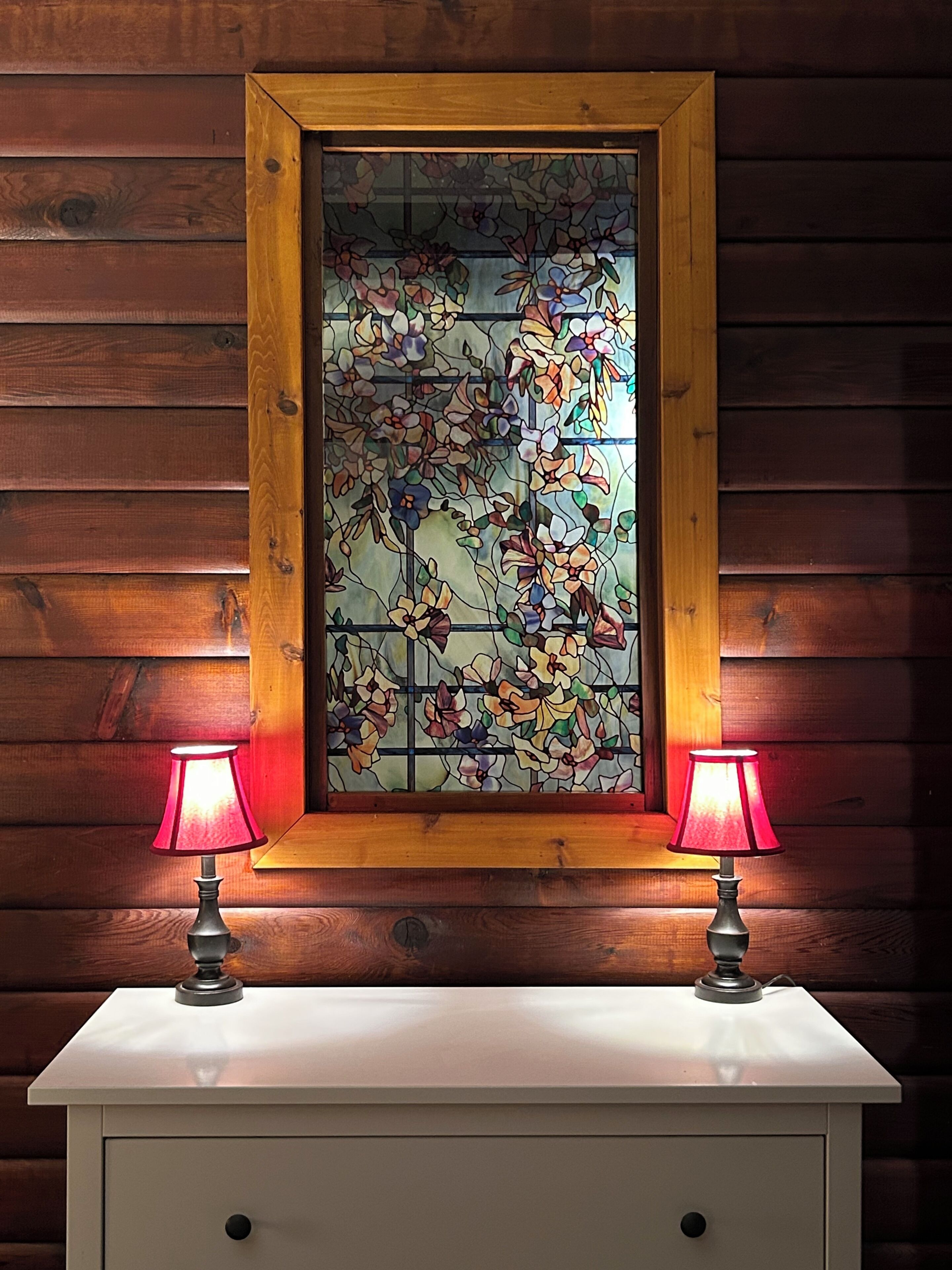 A wooden-framed stained glass window with floral patterns, flanked by two table lamps on a white cabinet.
