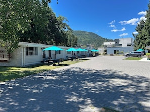 A row of mobile homes with outdoor seating and umbrellas, a paved driveway, and a mountain in the background. - The Cedars Motel (Sicamous)