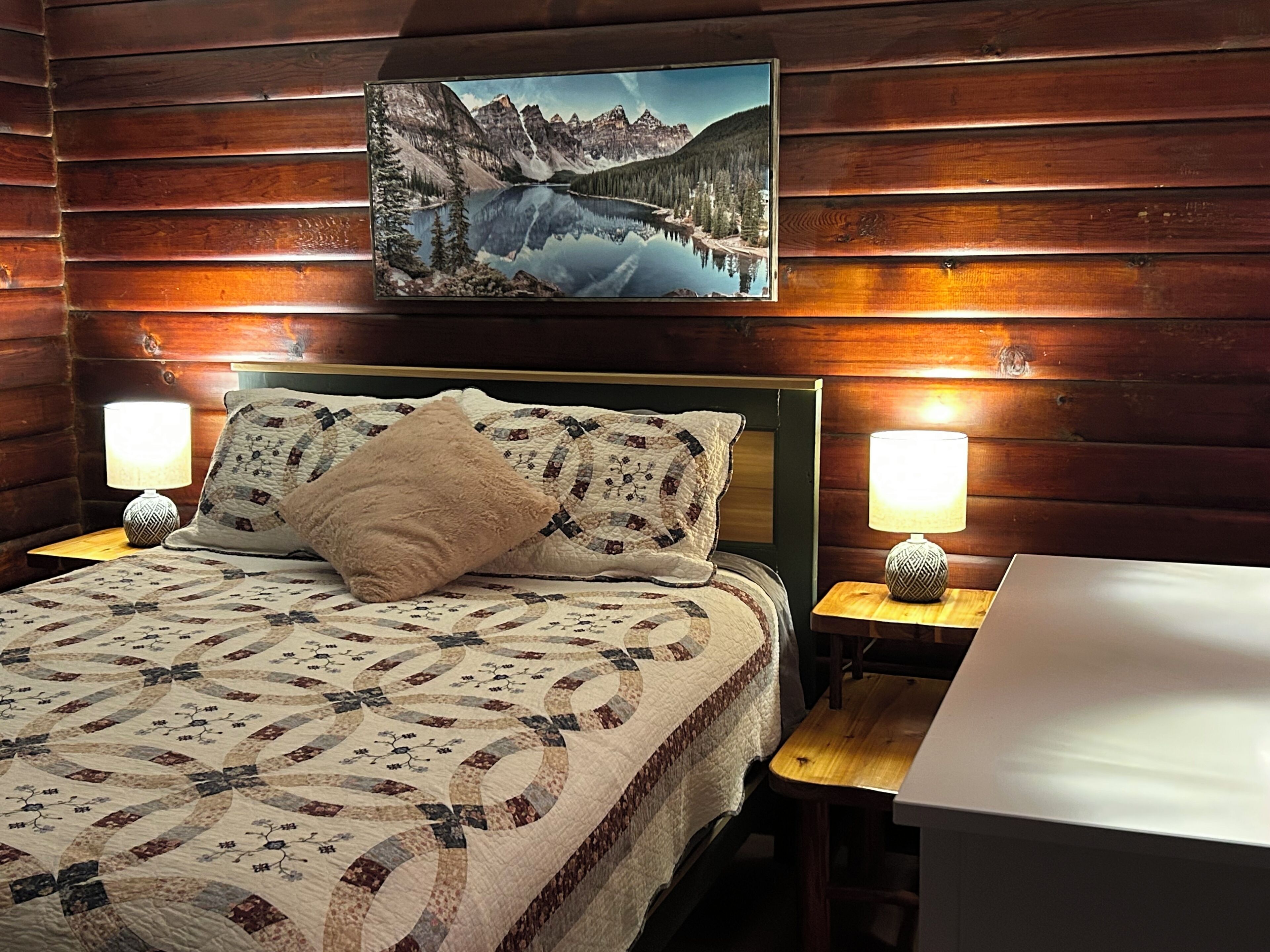 A wooden cabin room with a bed, two bedside lamps, a table, and a painting of a mountain lake on the wall.