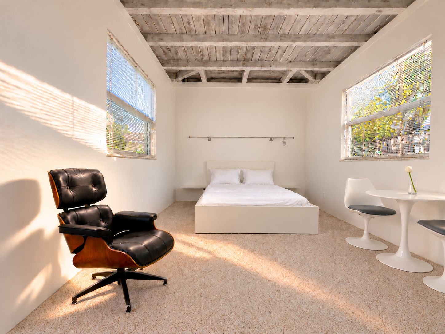 A bedroom with a bed, a chair, a small table, and two windows with blinds.