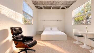 A bedroom with a bed, a chair, a small table, and two windows with blinds.
