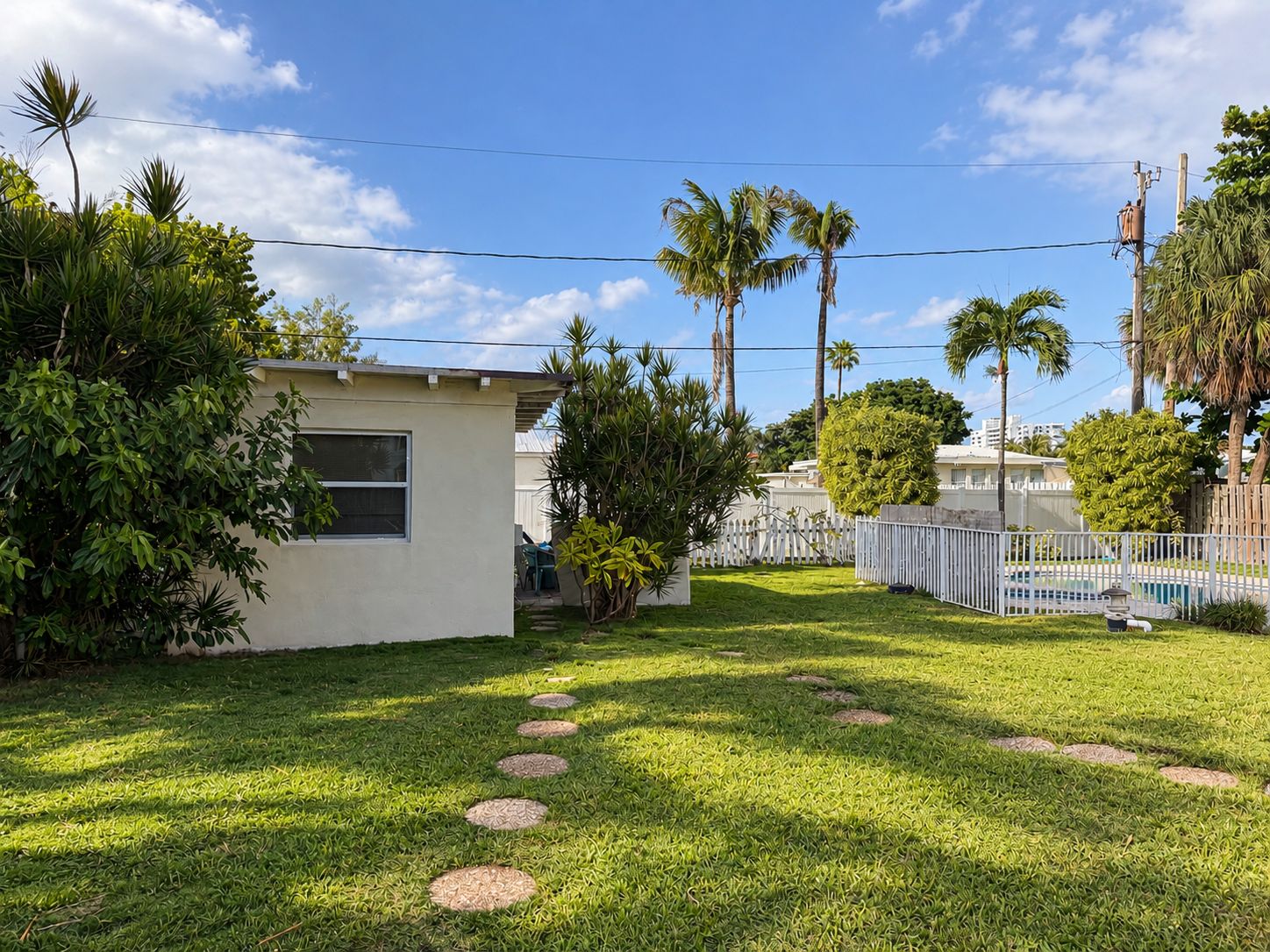 A residential backyard with a small house, palm trees, a white fence, and a swimming pool.