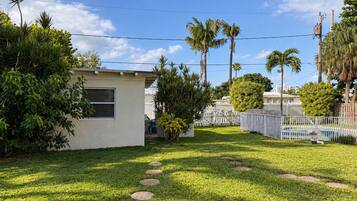 A residential backyard with a small house, palm trees, a white fence, and a swimming pool.