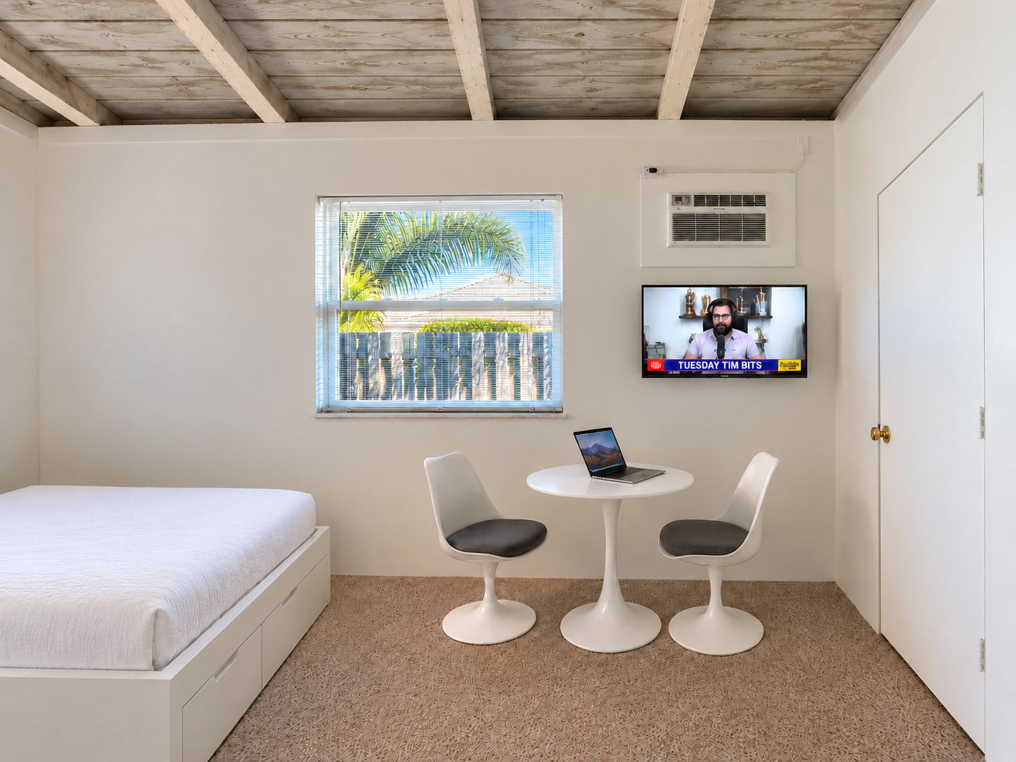 A room with a bed, two chairs, a table, a TV, and a window with a view of palm trees.