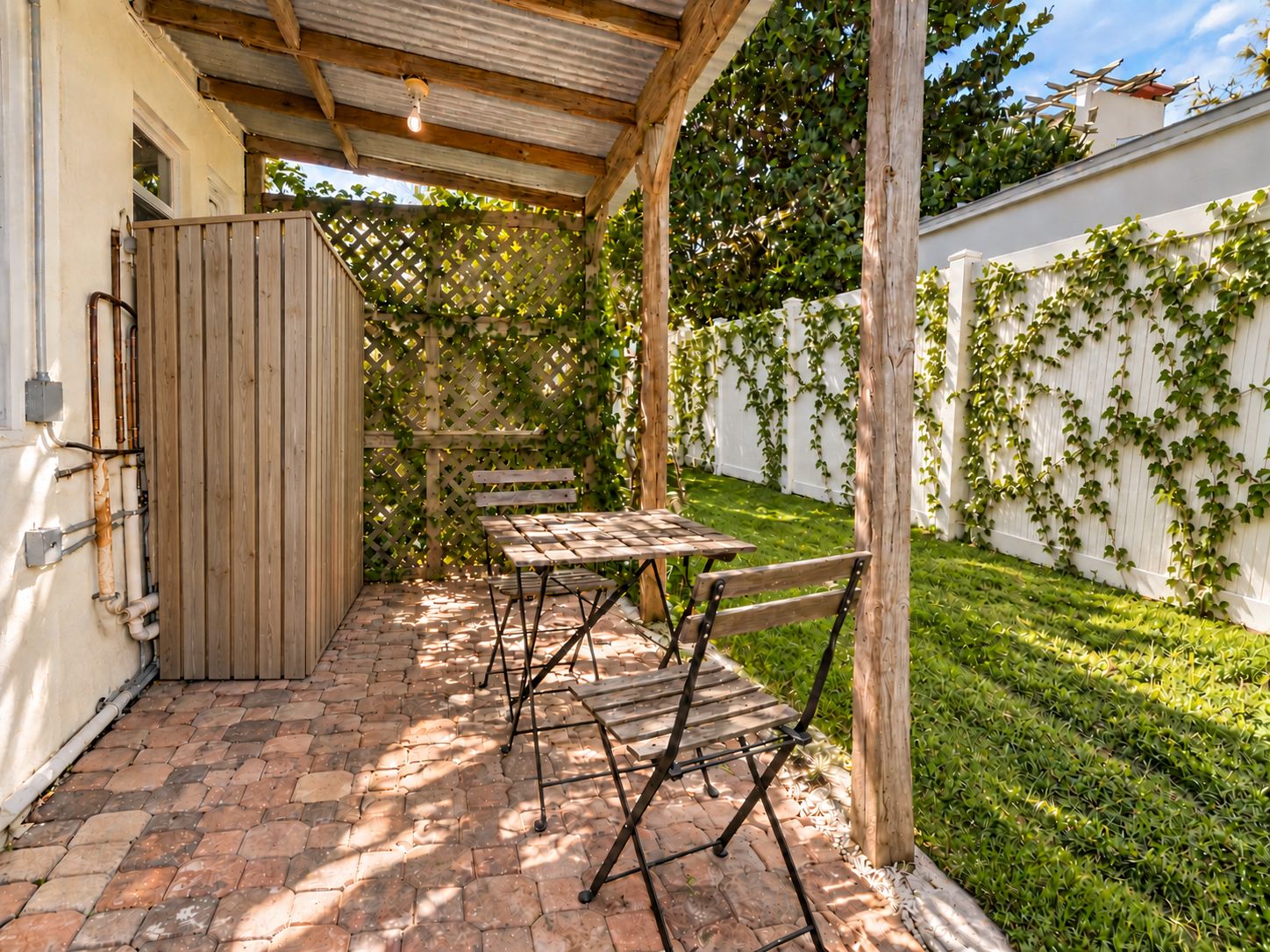 A covered patio with a wooden table and chairs, a wooden fence, and a white wall with greenery.