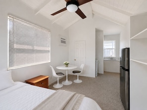 A bedroom with a bed, a bedside table, a chair, a small round table with a vase, a ceiling fan, a window with blinds, a door, and a refrigerator. - BEACHaus Cottage -Pool- 2 Blocks from Beach+Pier (Deerfield Beach)