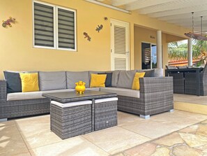 A sunlit outdoor seating area with a sofa and two side tables, a door with shutters, and decorative wall art. - Villa Paradise Cove – Your Gateway to Paradise (Jan Thiel)