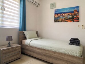 A single bed with a wooden frame, a bedside table with a lamp, a painting of a coastal cityscape, and a window with blinds. - Villa Paradise Cove – Your Gateway to Paradise (Jan Thiel)