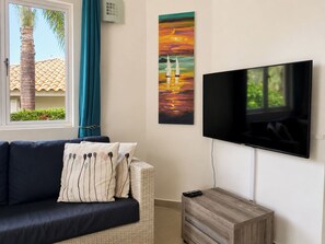A living room with a sofa, a flat-screen TV mounted on the wall, and a window with a view of a palm tree and building. - Villa Paradise Cove – Your Gateway to Paradise (Jan Thiel)